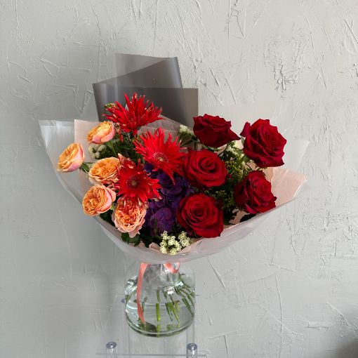 Elegant bouquet featuring red roses, orange and peach spray roses, red gerbera daisies, purple hydrangea, and assorted greenery, arranged in a clear glass vase.