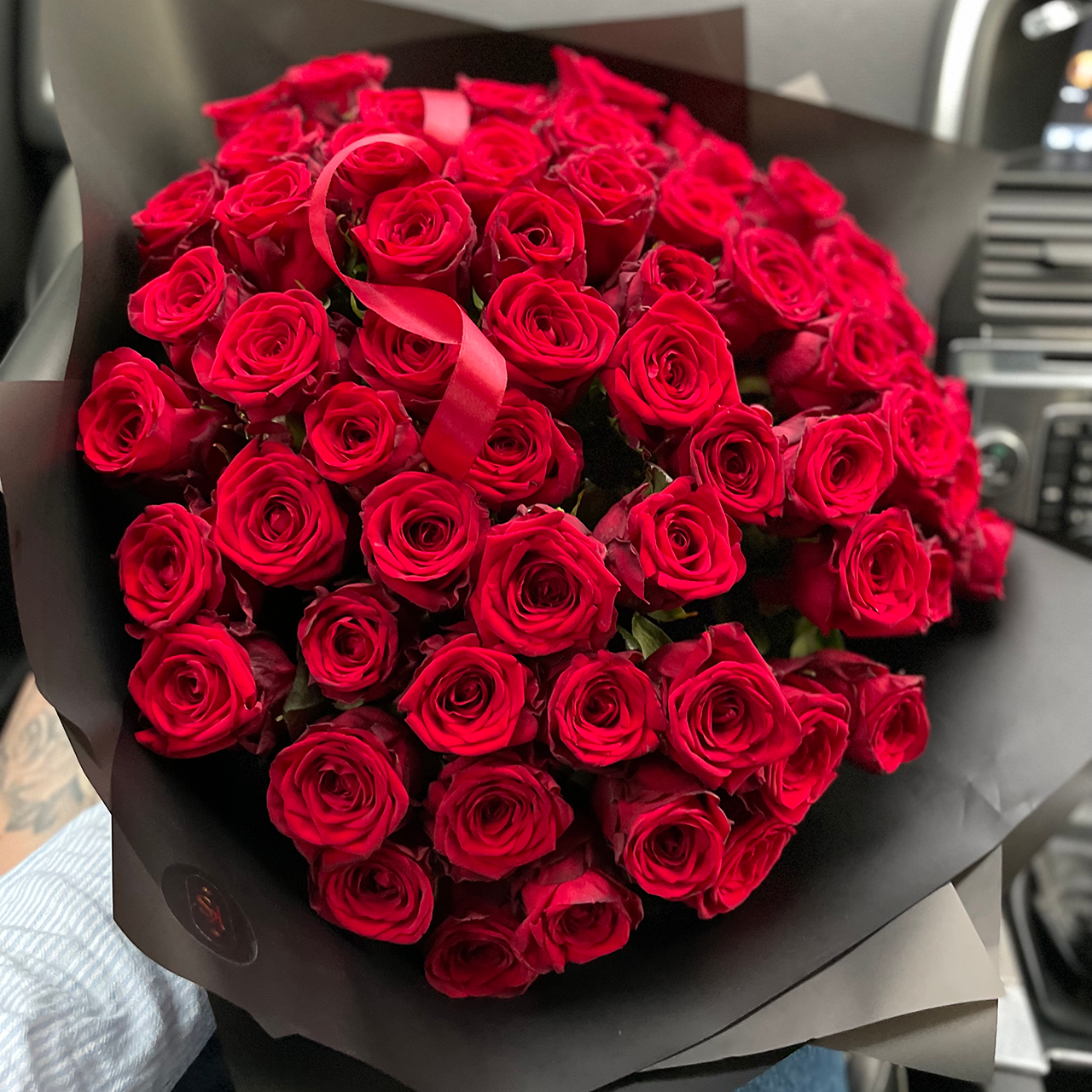 Lush bouquet of deep red roses wrapped in matte black paper with a red satin ribbon, held inside a car.