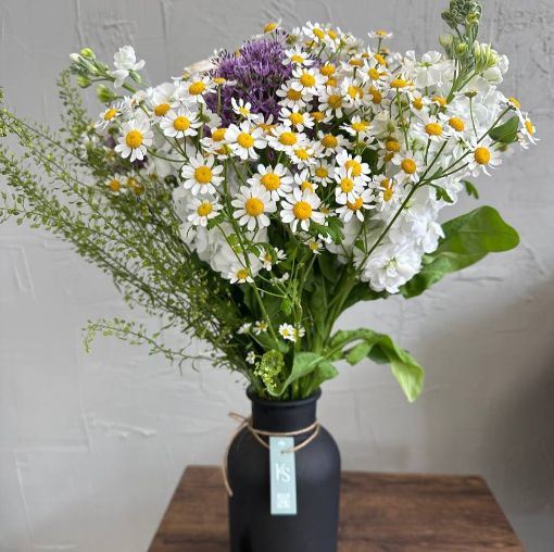 Fresh floral arrangement in a black vase featuring cheerful white chamomile daisies, white stock flowers, green foliage, and a hint of purple allium.