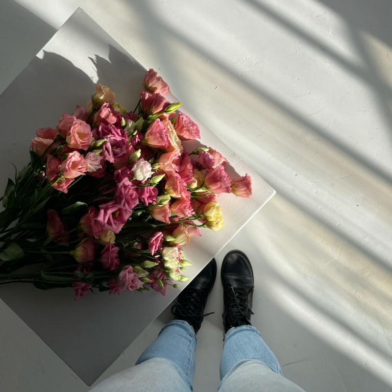 Bunch of multicoloured lisianthus flowers in soft pink, peach, cream and lavender tones resting on a light surface, captured from above with a person's feet in frame.