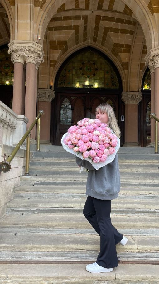 Florist carrying a large, lush bouquet of pink peonies wrapped in white paper, walking up the steps of a historic building.