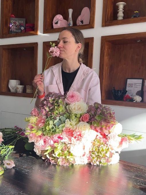 Florist inhaling the fragrance of a delicate pink flower, standing beside a large bouquet of roses, hydrangeas, and other soft pastel blooms in a bright flower shop.