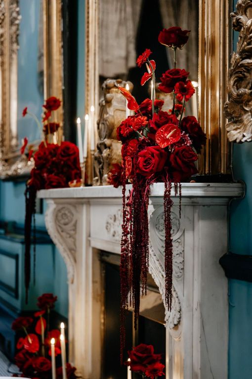 Opulent red rose and anthurium floral arrangement cascading down a marble fireplace, adorned with candles and surrounded by vintage mirrors in an elegant room setting.