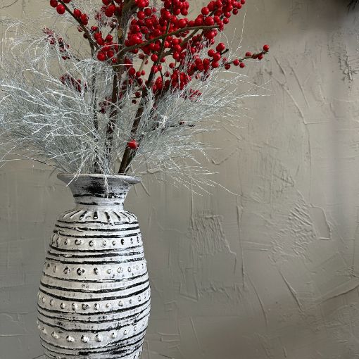 Decorative white vase with striped pattern holding sprigs of red berries and wispy silver foliage against a textured wall background.