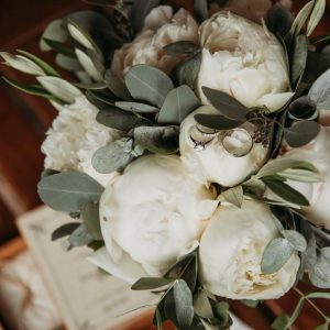 Close-up of a bridal bouquet with ivory peonies and eucalyptus, featuring a diamond engagement ring and wedding band.