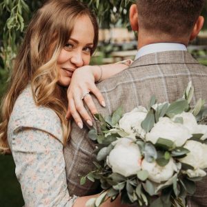 Bride embracing groom with white peony bouquet and engagement ring in focus.