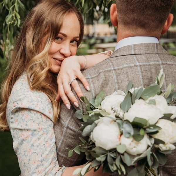 Bride embracing groom with white peony bouquet and engagement ring in focus.