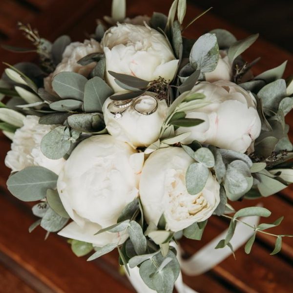 Elegant bridal bouquet of white peonies with eucalyptus foliage, adorned with wedding rings resting on a central bloom.