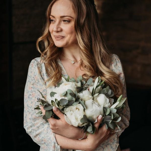Woman holding a bridal bouquet of white peonies and eucalyptus, smiling softly.