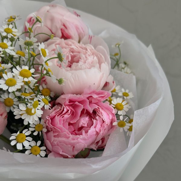 Close-up of a bouquet featuring soft pink peonies and delicate chamomile flowers wrapped in white paper.