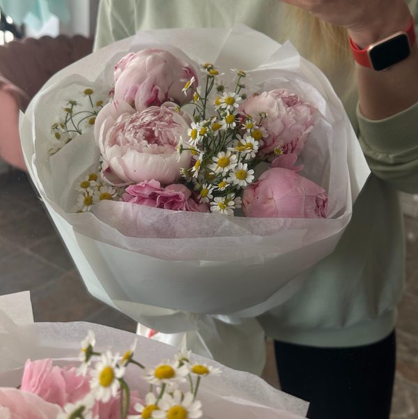 Woman holding a bouquet of pale pink peonies and chamomile flowers wrapped in white paper.