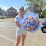 Woman holding a large summer bouquet of peach roses and blue hydrangeas, wrapped in KS Bloomery signature paper, outside a suburban home in the UK.