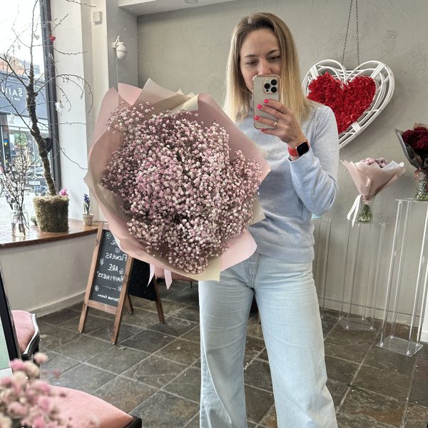Smiling woman holding a large bouquet of soft pink gypsophila flowers wrapped in pastel pink paper at a flower boutique.