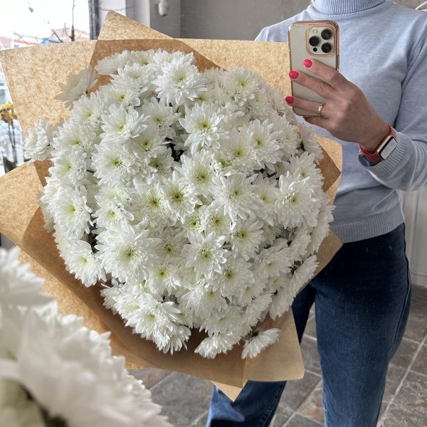 Large bouquet of white chrysanthemums wrapped in kraft paper, held by a person in a light blue jumper.