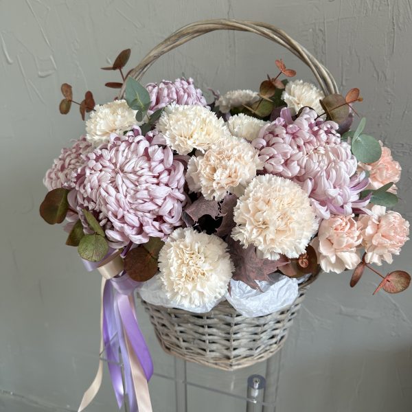 Pastel flower basket featuring light lilac chrysanthemums, creamy carnations, peach roses, and eucalyptus foliage, decorated with soft satin ribbons.