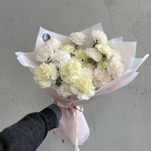 Elegant bouquet of cream and pale pink carnations wrapped in pastel pink paper, held in one hand against a textured grey background.