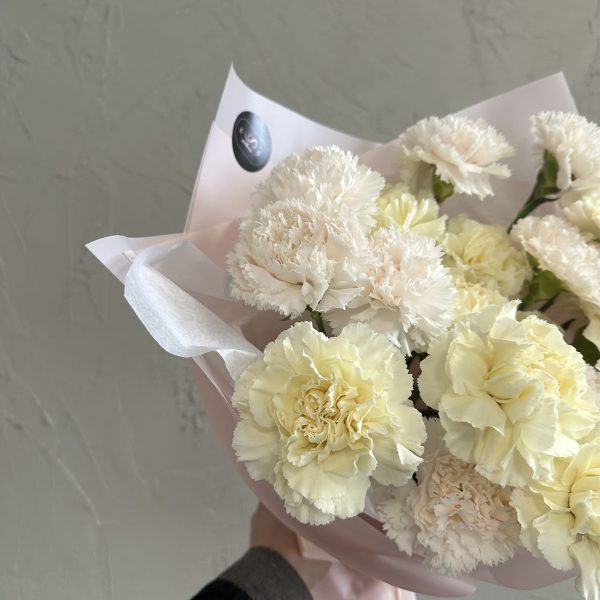 Close-up of a pastel bouquet with soft white and pale yellow carnations wrapped in pink tissue paper, held against a grey textured wall.