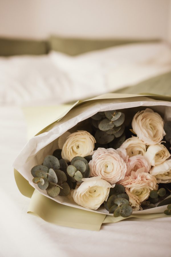 Pastel ranunculus and eucalyptus bouquet wrapped in sage green paper, resting on a white bed.