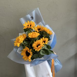 Bright sunflower bouquet wrapped in soft blue paper, held against a textured neutral background.