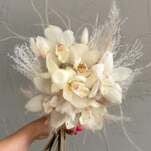 Ivory cymbidium orchid bouquet with pampas grass and dried florals, hand-held against a soft grey wall.