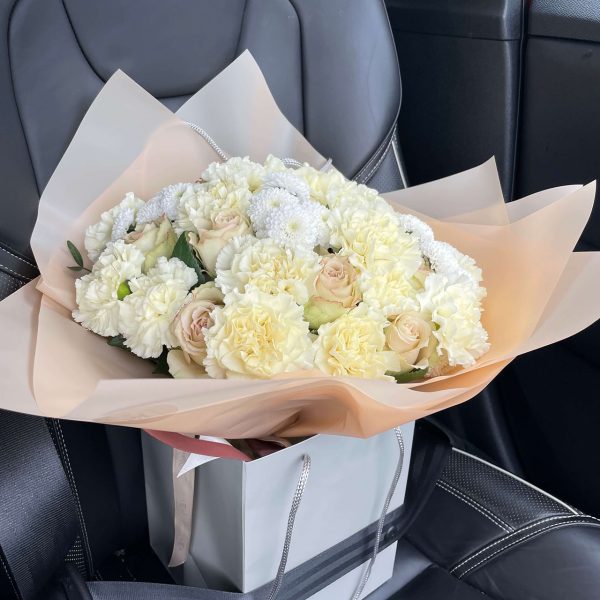Cream and white bouquet featuring carnations, roses, and chrysanthemums placed on a car seat in a stylish gift bag.