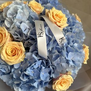 Close-up of a pastel bouquet with blue hydrangeas and peach roses, tied with a KS Bloomery branded ribbon.