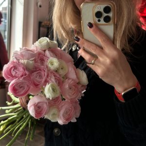 Woman holding a bouquet of soft pink and white ranunculus flowers while taking a mirror selfie.