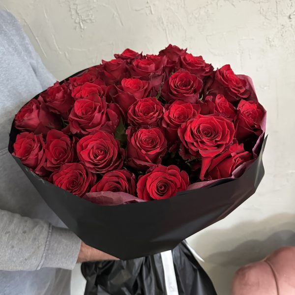 Large bouquet of classic red roses wrapped in black paper, held by a person in a grey jumper.