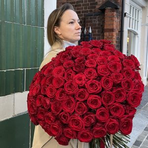 Woman holding a giant bouquet of red roses in front of a green tiled wall on a city street.