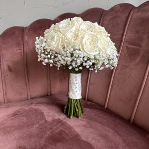 Classic white rose and gypsophila bridal bouquet with satin pearl wrap, displayed on a velvet pink sofa at KS Bloomery in Northampton.