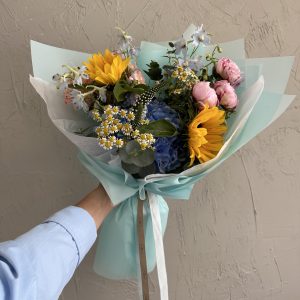 Hand-tied bouquet with sunflowers, chamomile, pink peonies, and blue hydrangeas in aqua wrapping.