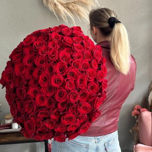 Woman holding an enormous round bouquet of red roses, wearing a red leather jacket and blue jeans.
