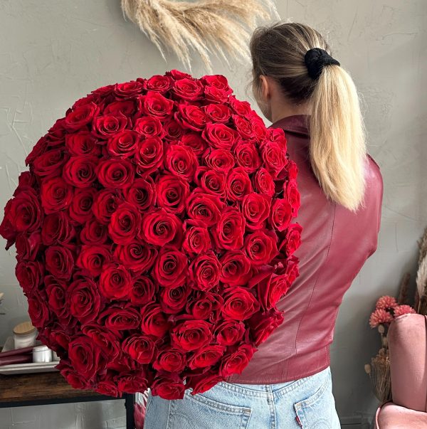 Woman holding an enormous round bouquet of red roses, wearing a red leather jacket and blue jeans.