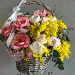 Wicker basket arrangement featuring yellow mimosa, coral roses, and soft pink ranunculus with white ribbon.