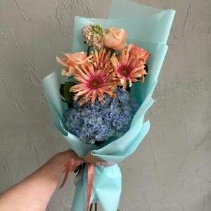 Delicate bouquet of peach gerberas, roses, and blue hydrangeas in turquoise wrap.