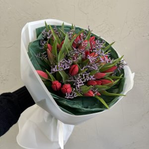 Bouquet of red tulips and delicate purple fillers, wrapped in white translucent paper with dark green tissue.