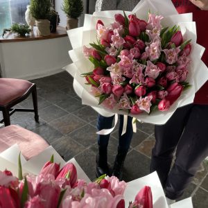 Large hand-tied bouquet of pink tulips and orchids wrapped in white paper, held indoors with morning sunlight