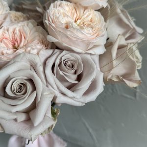 Close-up of a soft blush rose bridal bouquet with pale taupe roses and feathery grass accents, tied with a pastel pink ribbon.