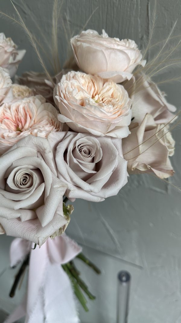 Close-up of a soft blush rose bridal bouquet with pale taupe roses and feathery grass accents, tied with a pastel pink ribbon.