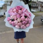 Large bouquet of fresh pink peonies wrapped in pastel blue paper, held outdoors on a residential street.