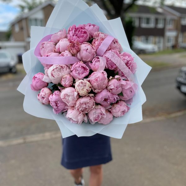 Large bouquet of fresh pink peonies wrapped in pastel blue paper, held outdoors on a residential street.