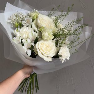 Elegant hand-tied bouquet featuring white roses, carnations, alstroemeria, and greenery wrapped in translucent paper.