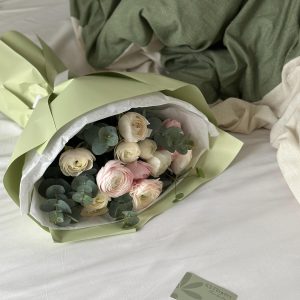 Elegant bouquet of pastel ranunculus and eucalyptus, wrapped in sage green paper and resting on a white bed.