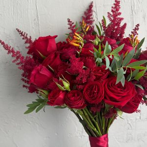 Close-up of a bridal bouquet with red roses, astilbe, green foliage and red ribbon wrap.
