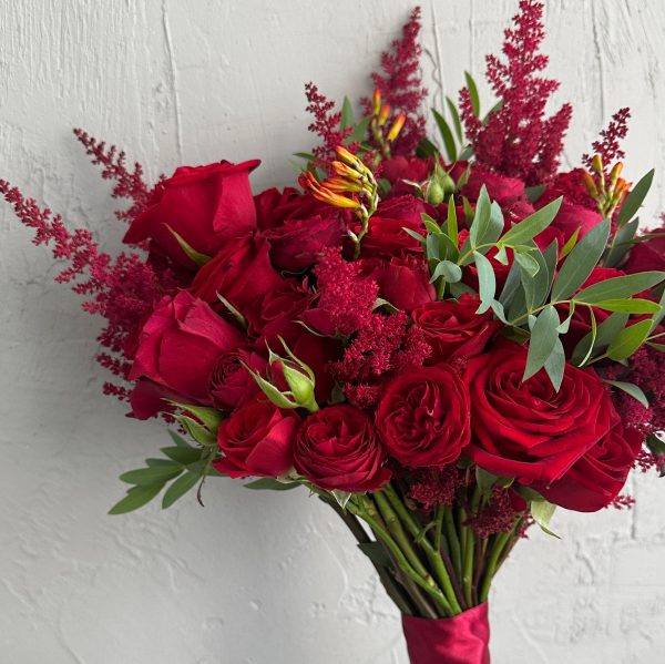 Close-up of a bridal bouquet with red roses, astilbe, green foliage and red ribbon wrap.