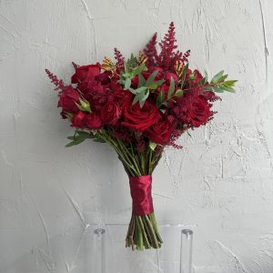 Red bridal bouquet with vibrant red roses, orange freesia, and burgundy astilbe, tied with satin ribbon, against a textured grey wall.
