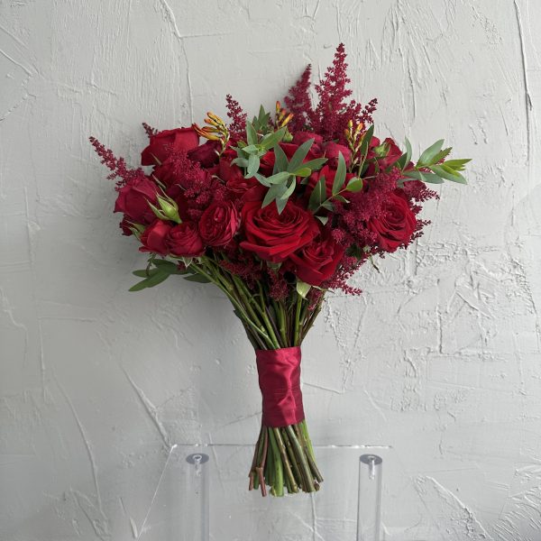 Red bridal bouquet with vibrant red roses, orange freesia, and burgundy astilbe, tied with satin ribbon, against a textured grey wall.