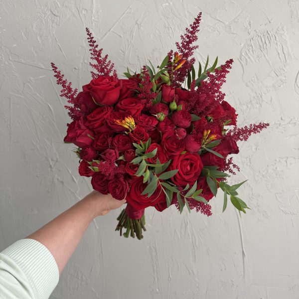 Hand-tied bouquet of red roses, astilbe, and green foliage held against a textured wall.
