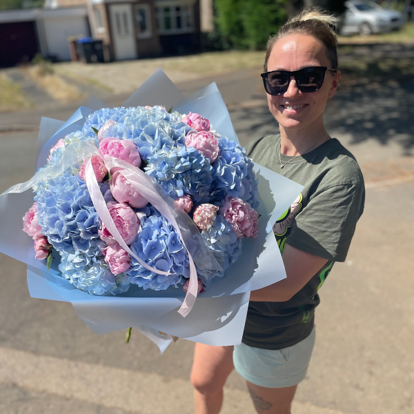 Smiling woman holding a large bouquet of blue hydrangeas and pink peonies wrapped in translucent paper with pastel ribbons.