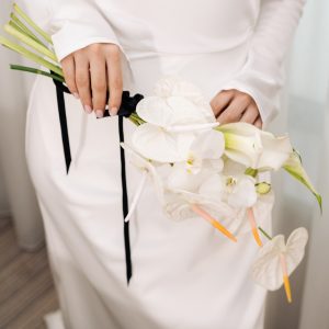 Close-up of a bride in a white gown holding a modern white bouquet with orchids, calla lilies, and anthuriums tied with a black ribbon.